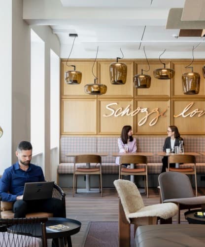 Colleagues in a café lounge with pendant lamps and wood-panelled booths, one working on a laptop.