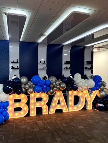 Illuminated BRADY marquee letters surrounded by balloons at a launch event.