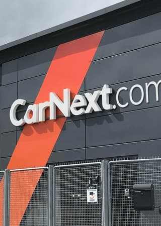The CarNext.com wordmark on a grey branch facade, the orange brand shape cutting across it behind a security fence.