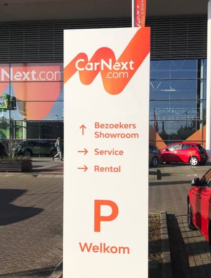 A CarNext directional totem at a site entrance, listing showroom, service and rental routes above a parking sign.