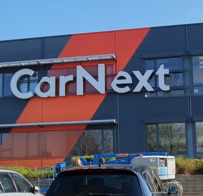 A CarNext branch by daylight, the orange brand shape cutting across its facade above parked cars.