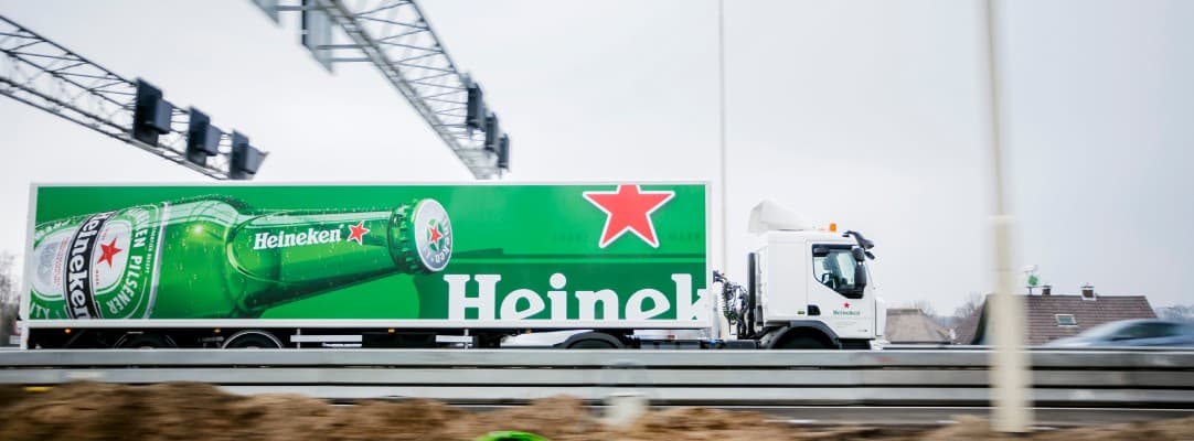 A Heineken articulated lorry with the green bottle graphic on its trailer, driving beneath a motorway gantry.