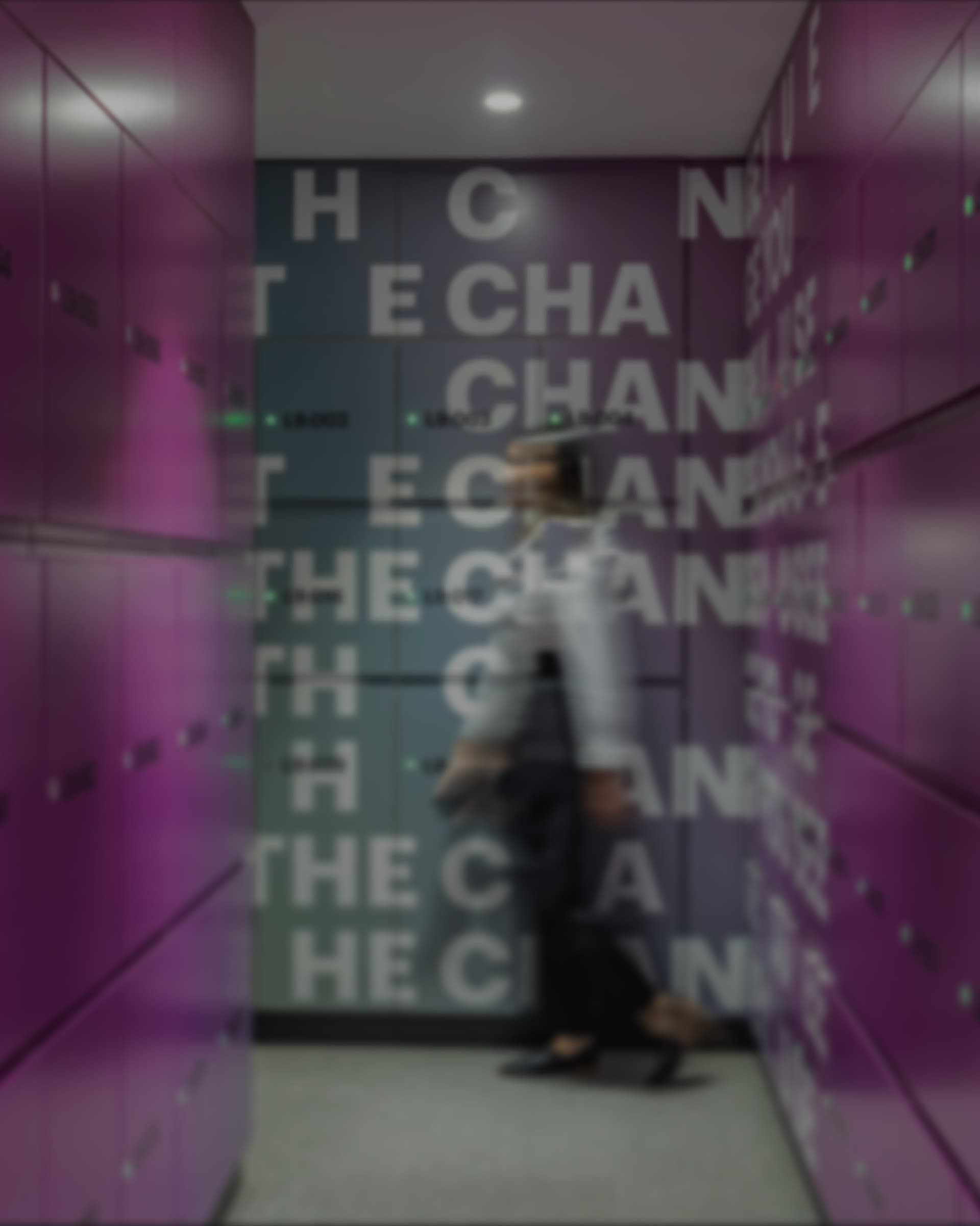 A locker bank corridor with magenta panels, wordmark lettering on the wall beyond, a colleague walking through.