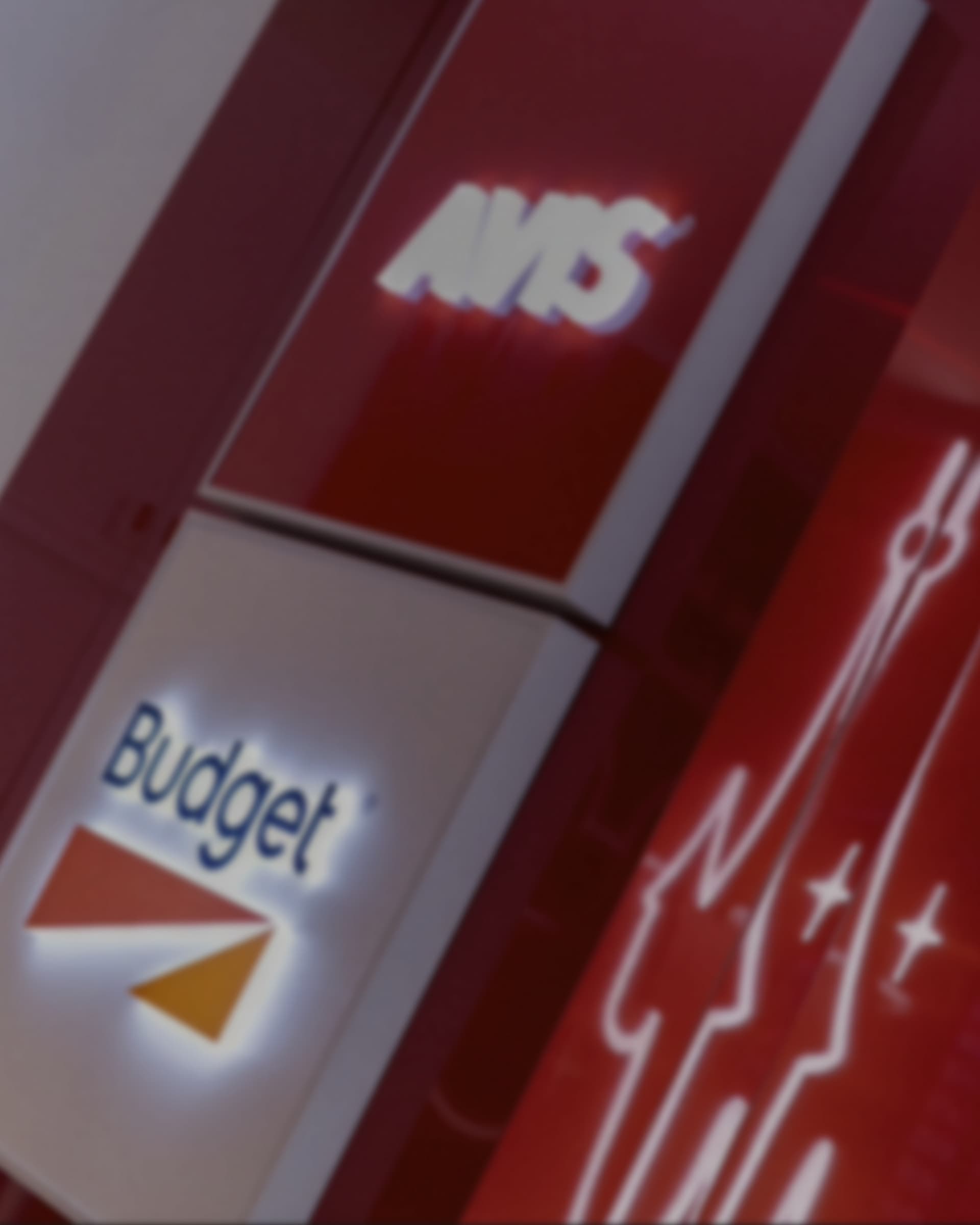 A close-up of illuminated Avis and Budget signs on a red gloss wall.