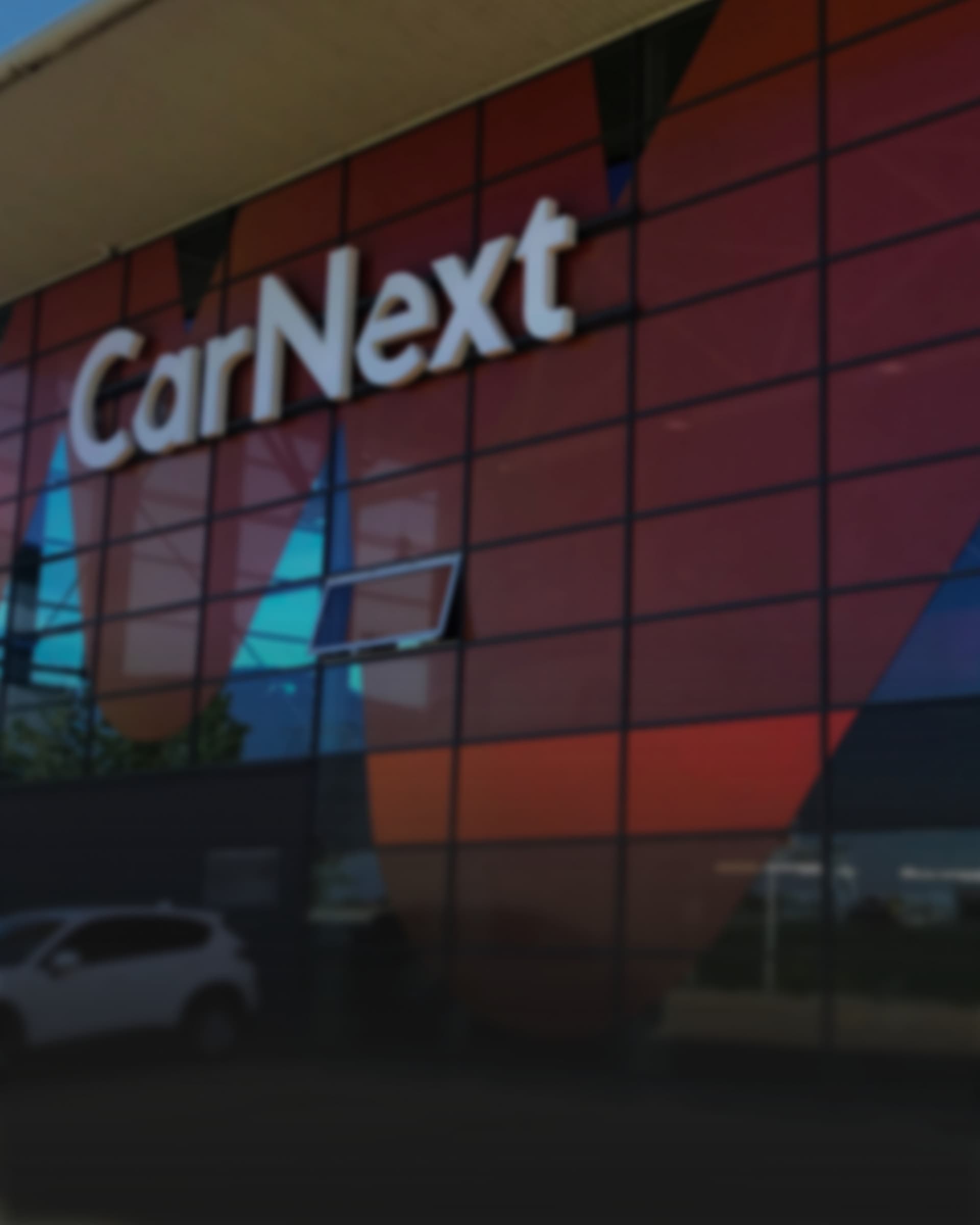A softened view of a CarNext branch's glazed facade at dusk, the wordmark lit above the entrance canopy.