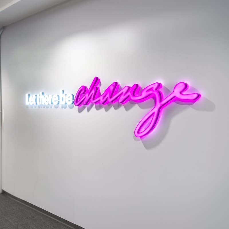 The words ‘let there be change’ in illuminated lettering on an office wall, the second half glowing magenta.