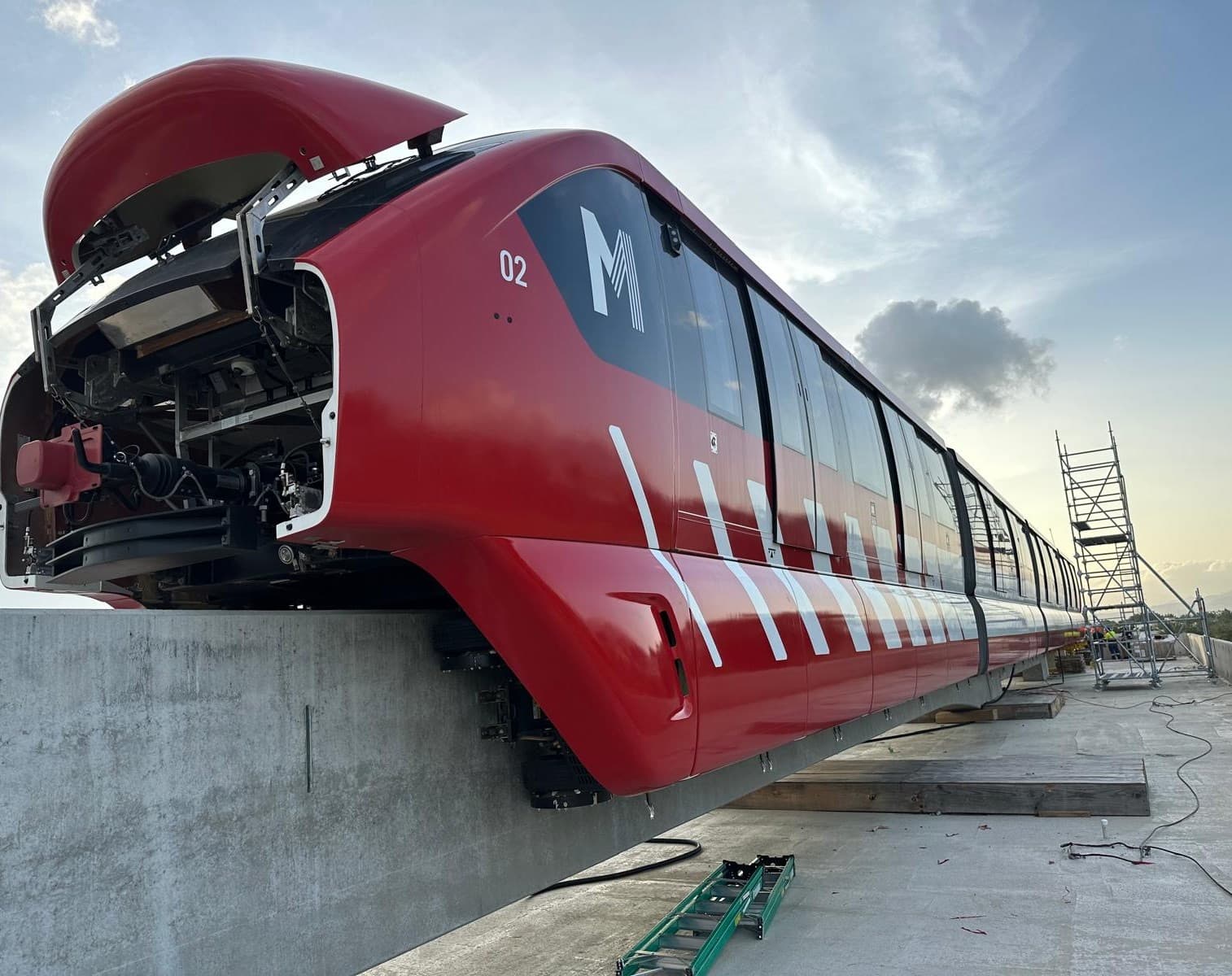 An Alstom metro car raised on a platform at the rail yard, its red and white livery applied along the full length of the body while the nose cone stands open for fit-out work.