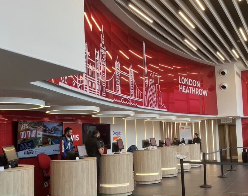 The Avis Budget counters at London Heathrow, a line-drawn skyline across the red wall.