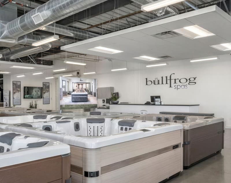 A Bullfrog Spas showroom floor lined with display spas, the wordmark on the back wall beneath exposed ceiling ductwork.
