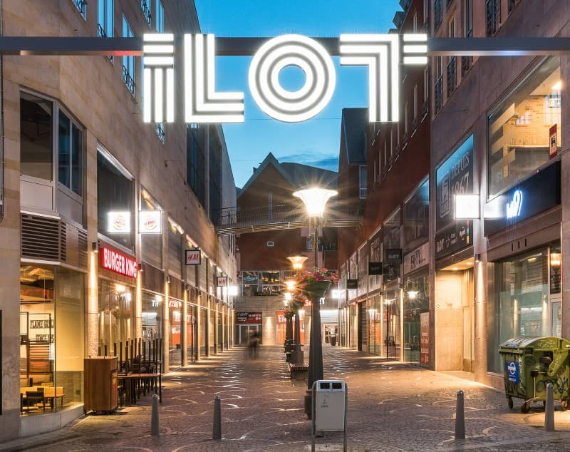 An illuminated ILOT sign spanning a street in the evening, storefronts lit below.