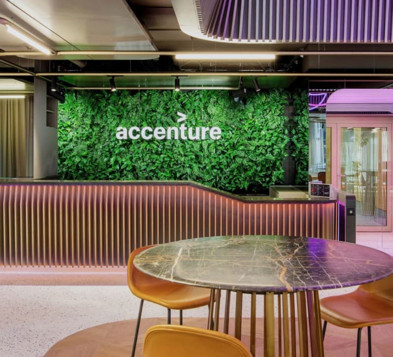 Accenture's wordmark on a green living wall behind a curved reception desk, under a purple neon ceiling installation.