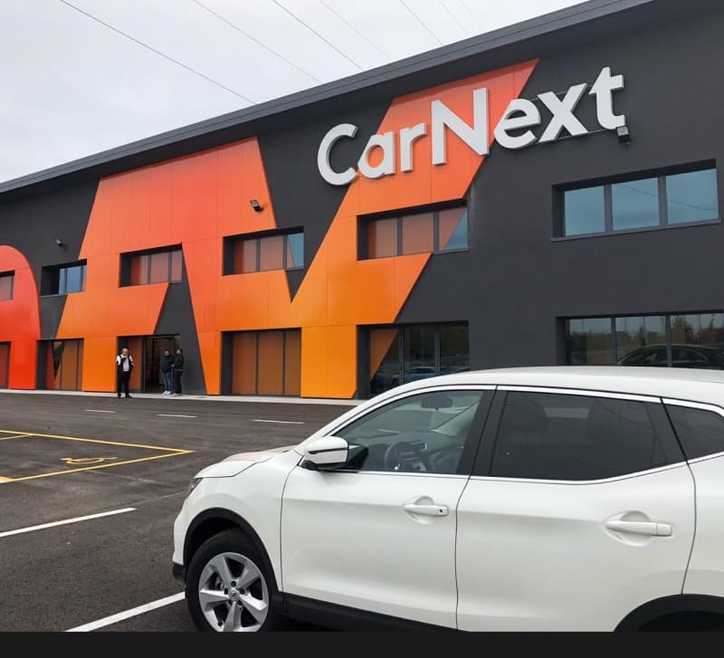 A CarNext branch facade, the orange brand shape cutting across it above a parked SUV.