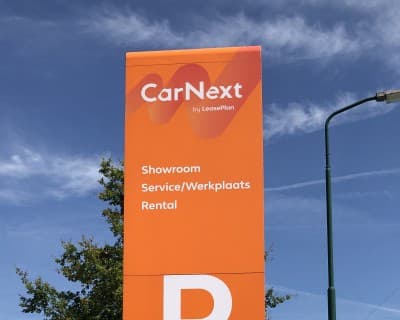 An orange CarNext directional totem sign against a blue sky at a branch entrance.