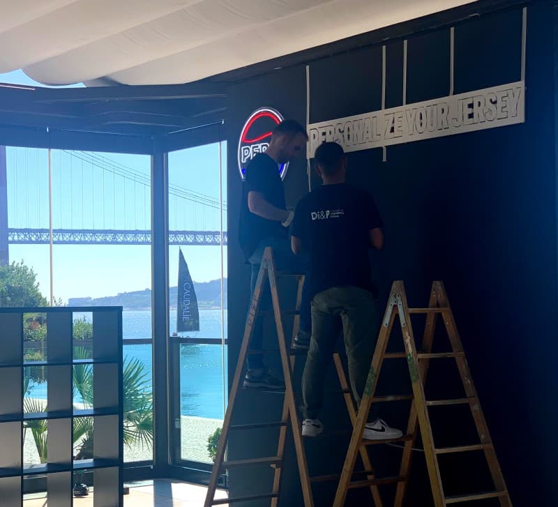Installers fitting an illuminated ‘Personalize your jersey’ sign to the activation’s wall, the Tagus and the 25 de Abril bridge visible through the glass behind it.