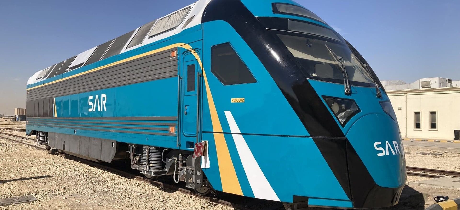 A Saudi Arabia Railways passenger train in blue, black and gold SAR livery, standing on desert track.