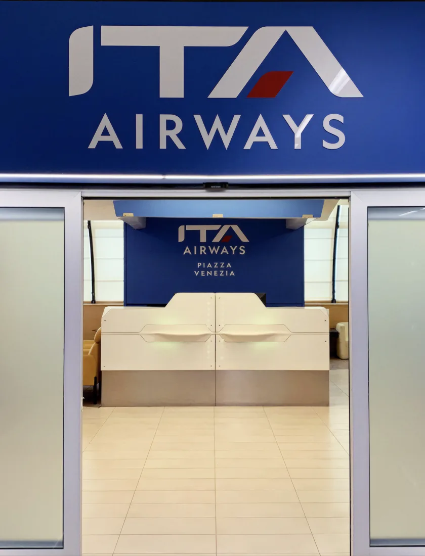 An ITA Airways lounge entrance inside an airport terminal, branded above the doorway and at the reception desk
